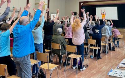 tuolijumppalaisia seisomassa jumpan vetäjän ohjaamana salissa