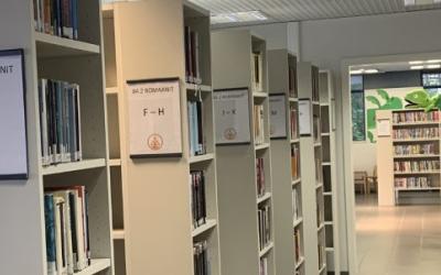 Kuva kirjaston sisäpuolelta. Kuvan vasemmalla reunalla on kirjahyllyjä, joiden kylteissä on kirjainopasteet.