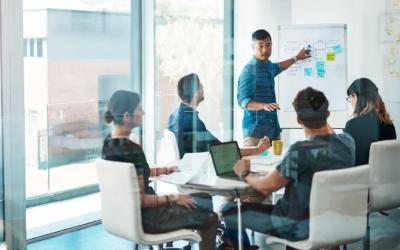 neljä ihmistä istuu pöydän äärellä ja yksi ihminen seisoo esitystaulun äärellä