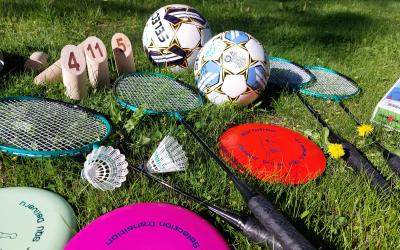 Kuvassa on vihreälle nurmikolle aseteltuja liikuntavälineitä. Kaksi jalkapalloa, neljä sulkapallomailaa, kaksi sulkapalloa, kolme frisbeetä, ja mölkyn pelinappuloita.