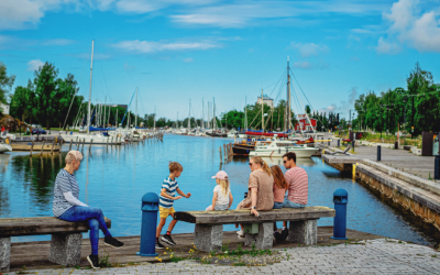 2 aikuista ja 3 lasta istuvat Uudenkaupungin kaupunginlahden rannalla penkillä. Taustalla sininen taivas, merta ja veneitä.