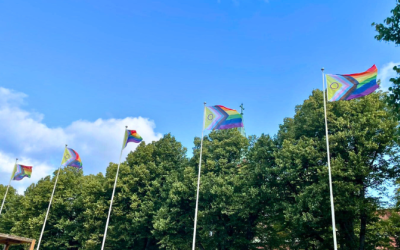 pride-liput liehuvat kesäisessä säässä viidessä lipputangossa. Taustalla taivasta ja vihreitä puita.