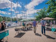 Kaksi ihmistä kävelee kaupunginlahden rannalla kesäisenä päivänä. Taivas on hieman pilvinen, vedessä veneitä.