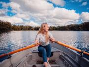 Vaaleatukkainen tyttö soutaa järvellä valkoinen paita päällä. Taivas on sininen ja hieman pilvinen.