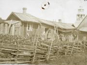 Baltzarborg. Pieni huonokuntoisen näköinen puinen asuinrakennus, ns. Lindgrenin mökki. Etualalla riukuaita, kasvimaa, kaksi naista rakennuksen edustalla, taustalla vanhan kirkon torni.