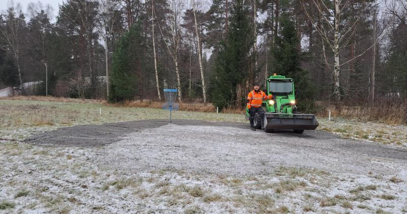Pellolla frisbeegolfkori ja mies traktorin kanssa taustalla metsää