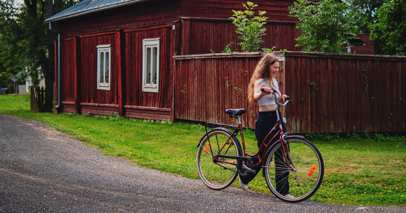Nuori nainen kävelee ja taluttaa samalla pyörää. Taustalla punainen vanha rakennus.