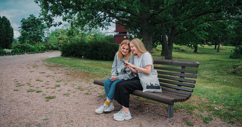Kaksi tyttöä istuu puistonpenkillä ja katsovat puhelinta.