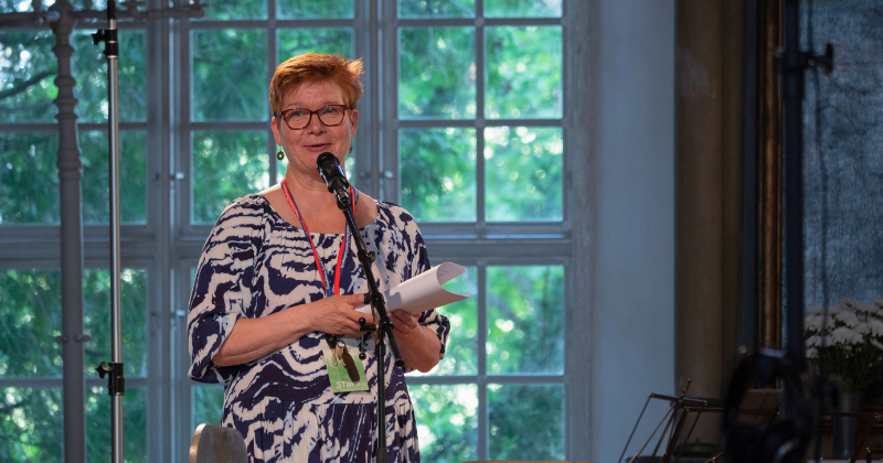 Nainen pitää puhetta, taustalla vanhan kirkon iso ikkuna ja vehreän kesäinen maisema.