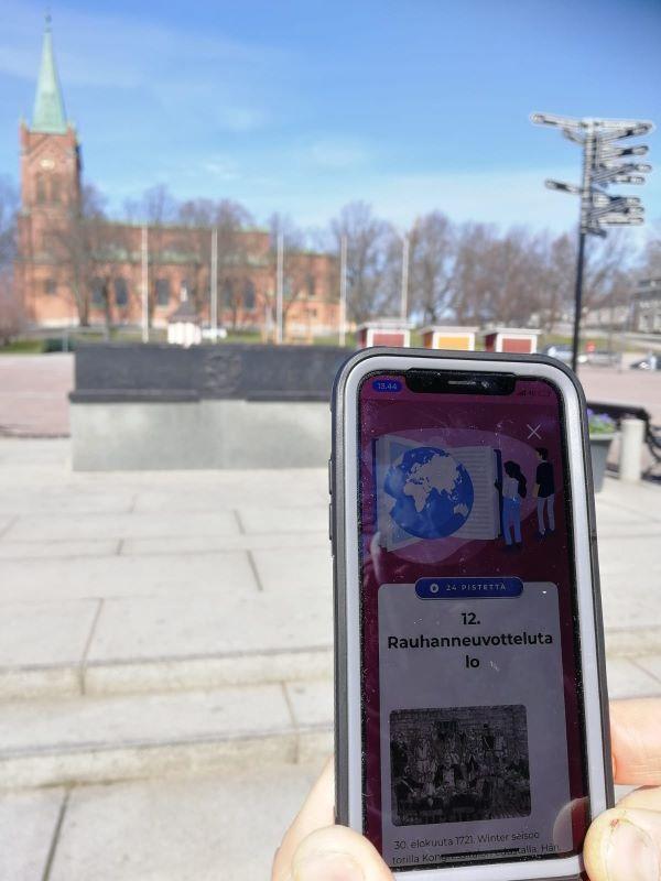 Puhelimen näyttö, jonka takana näkyy tori ja kirkko.