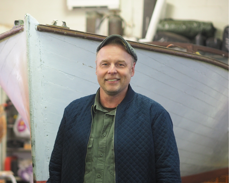 Asmo Hällfors seisoo puuveneen edessä, hymyilee ja katsoo kohti.