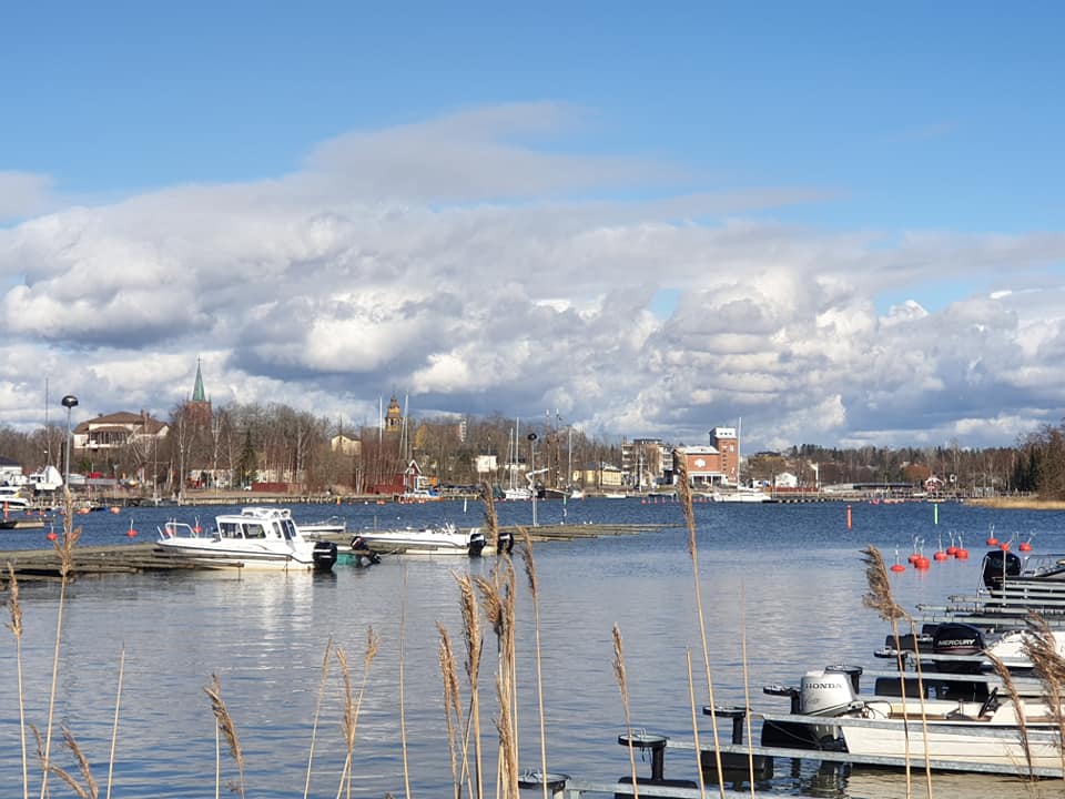Kaunis, huhtikuinen kuva Kaupunginlahden alkupäästä, jossa näkyy edessä kaisloja, niiden takana laitureissaan olevia veneitä, ja taaempana näkyy kaupunki.